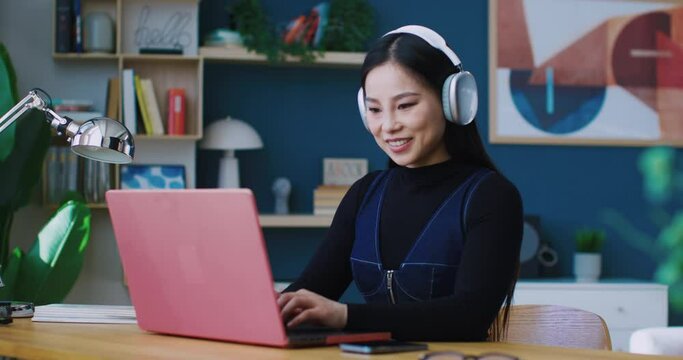 Beautiful Asian woman smiling to herself while working remotely. Listening to music in large white headphones. Enjoying good quality of sound. Finding ways to making work more relaxing.