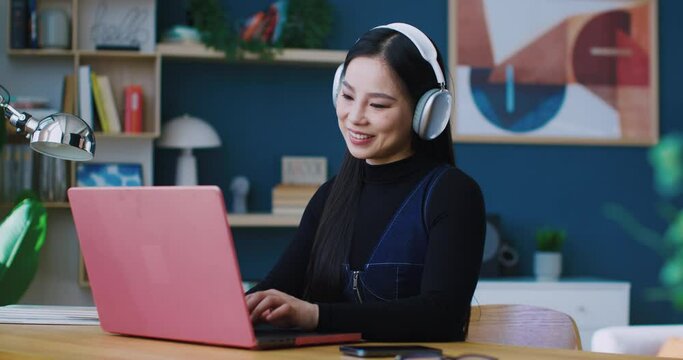 Charming Chinese girl teaching or studying remotely while listening to music. Getting distracted by beats playing from large white headphones. Beginning to nod her head to beat in song.