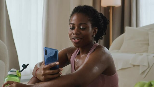 young black woman doing a workout at home, takes a break to rest and drink water, skin is wet. Uses mobile phone to open app for online training and chat. Smiles and feels contented and pleased