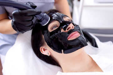Close-up of a young woman in a black cosmetic mask at a spa procedure. A cosmetologist performs a...