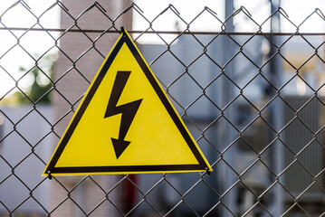 Close-up Of A Yellow High Voltage Caution Sign Hanging On Chain Fence. Shallow Depth Of Field