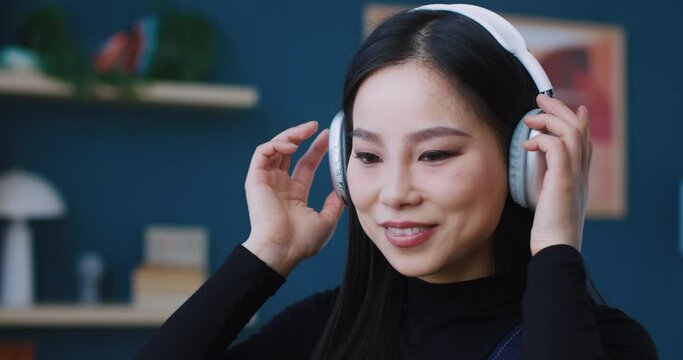 Portrait of cute Asian girls face. Woman putting on brand new headphones. Enjoying great sound quality. Listening to music and nodding head to beat in favourite song. Working or studying.