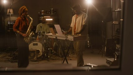 Full handheld, shot from control room window, of African American blues male and female band members playing saxophone, keyboards and drums, while performing together in recording studio