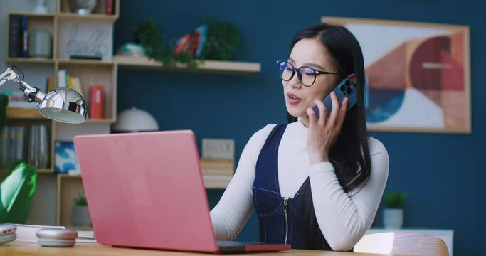 Chinese girl with big glasses sitting at desk in modern apartment. Picking up phone from table. Having very fast conversation with someone. Focused on studying or working. Using laptop.
