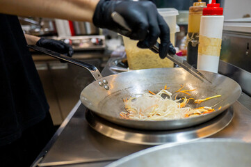 A chef in black gloves stir-fries noodles with vegetables in a stainless steel pan in a professional kitchen