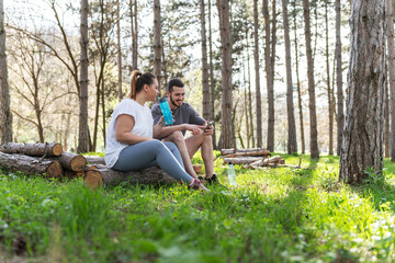 Sitting on a fallen tree trunk, the couple, including the determined overweight female, catch their breath, their laughter blending with the rustling leaves as they enjoy a post-workout moment.	