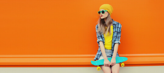 Summer portrait of stylish young woman with skateboard in colorful clothes on orange background