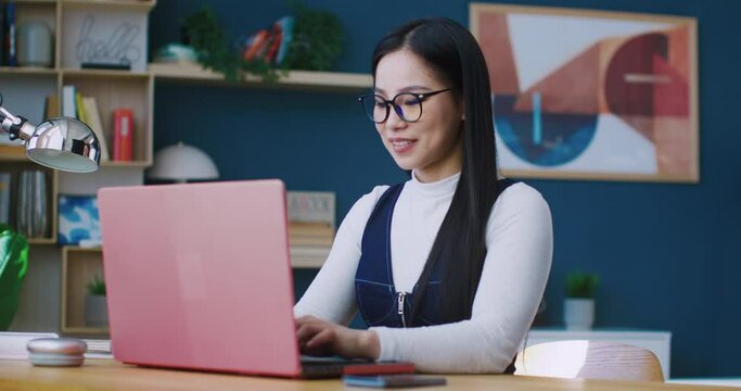 Beautiful Asian woman sitting at her desk while working or studying on laptop. Pressing keys on keyboard. Looking directly at camera and beginning smiling. Showing heart symbol with two fingers.