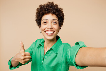 Close up young woman of African American ethnicity wear green shirt casual clothes doing selfie shot pov on mobile cell phone show thumb up isolated on plain pastel beige background Lifestyle concept