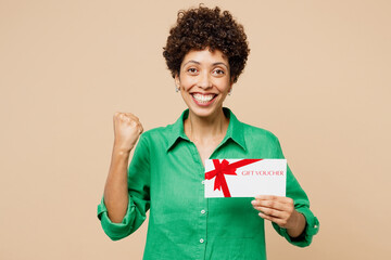 Young woman of African American ethnicity wear green shirt casual clothes hold gift certificate coupon voucher card for store do winner gesture isolated on plain beige background. Lifestyle concept.