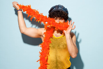 Young happy gay Latin man wear mesh tank top hat yellow clothes hold in hand cover mouth with tinsel isolated on plain pastel light blue cyan background studio. Pride day June month love LGBT concept.