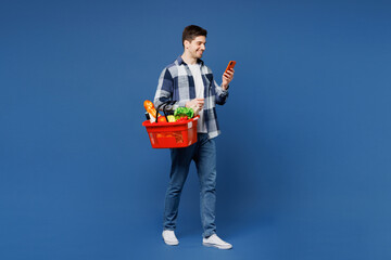 Full body young man in shirt white t-shirt casual clothes hold basket bag for takeaway with food products use mobile cell phone isolated on plain blue background Delivery service from shop restaurant