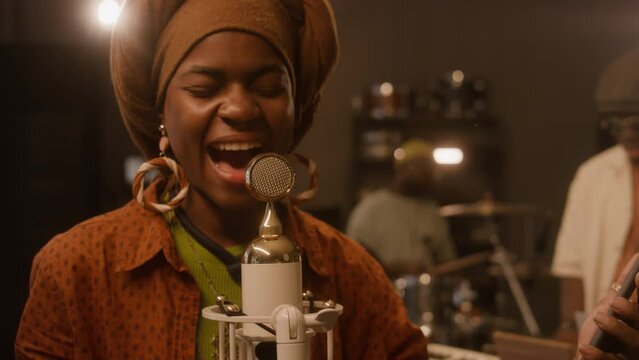 Medium close-up of emotional African American female jazz musician in ethnic turban singing into vintage microphone while performing with band at gig in nightclub, filming herself on smartphone