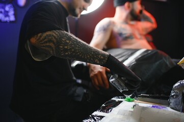 bearded tattoo artist working at his studio tattooing sleeve on the arm of his male client. Man...