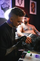 bearded tattoo artist working at his studio tattooing sleeve on the arm of his male client. Man getting tattooed by professional tattooist