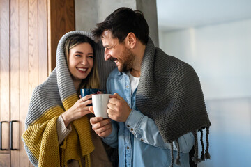 Happy young couple with hot drinks cuddling under warm blanket at home