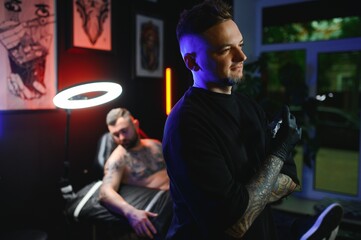 tattooer in black gloves dreamily looking in camera holding tattoo machine in hand in modern tattoo studio