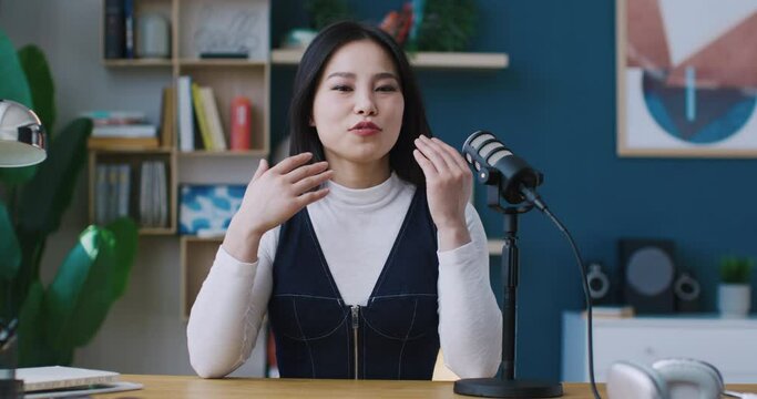 Asian girl looking directly at camera. Beginning to talk and move hands. Greeting by waving. Saying hello. Microphone setup next to female. Online class or conversation. Heart symbol with two fingers.