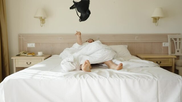 A woman on a white bed throws a black bra towards the camera at me from under the covers. Bright hotel room with early morning sunlight.