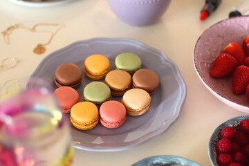 Plate of pastel macarons, cookies and chocolate, cup of tea of coffee, glass of bubble water, various berries, books and accessories on the table. Selective focus, pastel colors.