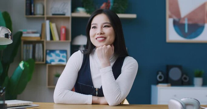 Camera closing in on Asian female. Girl sitting in front of desk. Beginning dreaming of something. Afterwards looking directly at camera while smiling. Placing hand under her chin.