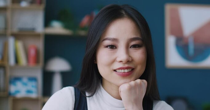 Portrait of Asian woman with brunette hair glancing directly at camera. Cheerfully smiling and having good feelings during rest at home. Relaxation alone. Beautiful face. Happiness concept.