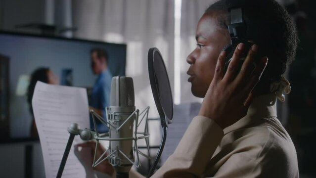 Medium side close-up of African American female voice-over actress in headphones reading dialog lines from cue sheet into microphone, while doing movie dubbing work in recording studio