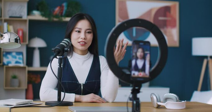 Cute Asian blogger using modern technology devices for recording video on smartphone. Streamer telling interesting story in microphone while looking at camera. Phone in holder with circular lights.