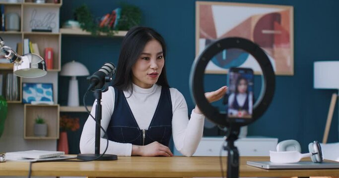Famous Chinese vlogger or streamer speaking into microphone with her followers live on phone. Smartphone in middle of holder with circular lights. After conversation showing heart with her hand.