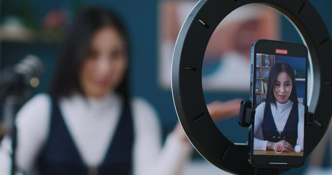 Camera focused of smartphone held by stand with circular lamp. Video recording. Young woman telling something in microphone or filming advertisement on phone. Showing gesture of love with hand.