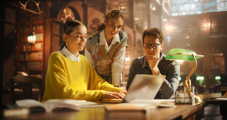 Engaged Group of Students Collaborating Over a Laptop in a Cozy Library Setting, Surrounded by Books. Two Women and One Man Discussing Academic Project, Focused and Smiling.