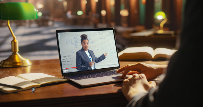 Student Learning From Online Lecture on Laptop in Library. African American Female Professor Teaching Engineering, Displayed on Screen, With Books and Notebook on Wooden Table