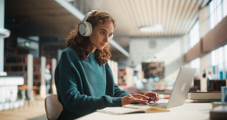 Concentrated Young Woman Studying in Modern Library, Wearing Headphones and Using Laptop for...