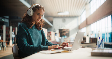 Concentrated Young Woman Studying in Modern Library, Wearing Headphones and Using Laptop. Female...
