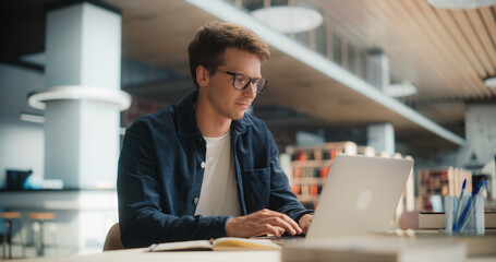 Concentrated Young Caucasian Male Student Working on His Laptop in a Modern Library. Man Engaged in...