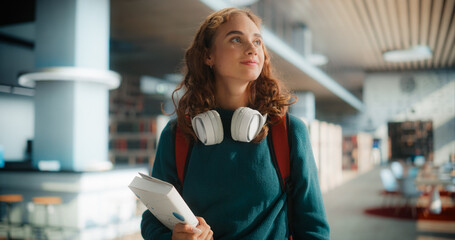 Dreamy Young Female Student Wandering Through Modern Library With Books and Headphones, Wearing...