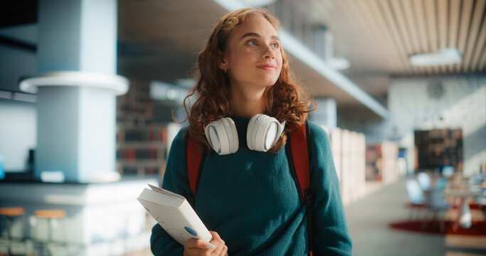Dreamy Young Female Student Wandering Through Modern Library With Books and Headphones, Wearing Casual Green Sweater and Carrying Red Backpack, Looking Upward With Curious Expression