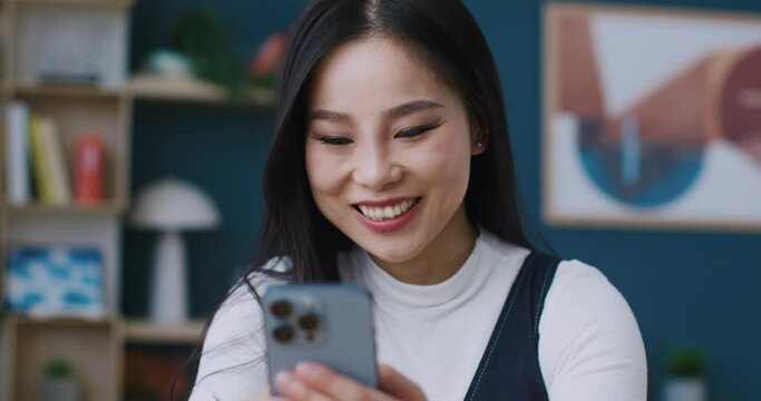 Beautiful Asian woman using her personal phone for remote communication from home. Satisfied young girl typing fast response or message to her boyfriend on smartphone. Positive online chat.