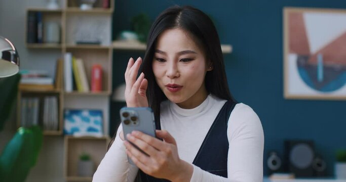 Charming Asian woman looking surprised after reading funny message or good news on mobile phone at home. Young female positively smiling while using smartphone. Chatting online with someone.