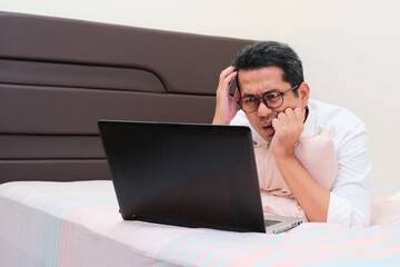 A man on bed showing confused face expression when looking to his laptop