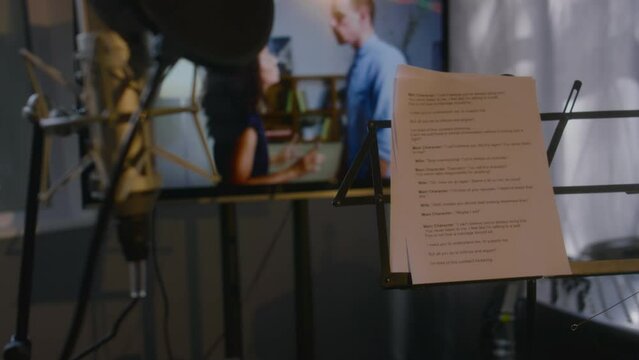 No people medium close-up of workplace of voice-over actor, with studio microphone, pop filter, stand with script and dialog cue sheets, and movie scene playing on screen of diverse couple fighting