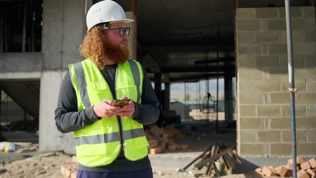 Construction worker with red beard in glasses and safety helmet procrastinates on internet during a break, surfs site in browser, scrolls social networks stand outdoors construction site.