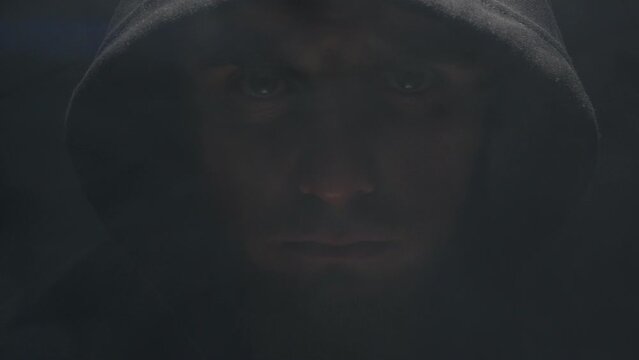 a hooded man in the dark raises his head and looks at the camera. evil man