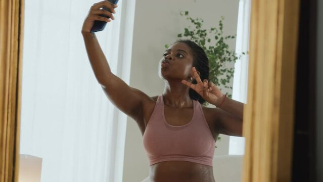 Young black woman doing a workout at home, takes a break to rest and uses a mobile phone. Uses online app for training and chatting. Smiles and feels contented and pleased, taking selphie photo