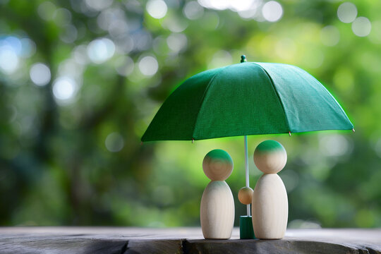 Green umbrella sheltering couple of wooden figurines, set against blurred natural backdrop, protection or insurance. Security and care, concepts related to safety or family protection in advertising.