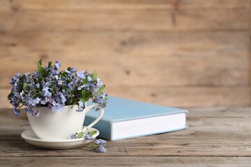 Beautiful forget-me-not flowers in cup, saucer and book on wooden table, closeup. Space for text