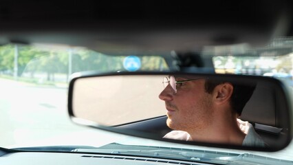 Happy young male driver in glasses carefully looking at the road in the city and looking in the rear view mirror. Transport and lifestyle concept. Real time