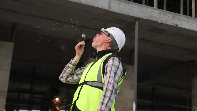 Charismatic engineer builder blows dandelion while standing against the background of welders of an unfinished building with scaffolding, break at works, slow motion.