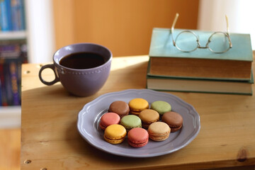 Purple plate filled with pastel macarons, cup of tea or coffee, vintage books and reading glasses on the table. Colorful bookcase in the background. Selective focus.