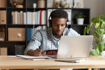 Focused young smart mixed race business man wear headphones while learning language by watching webinar on laptop and write notes during online courses lessons